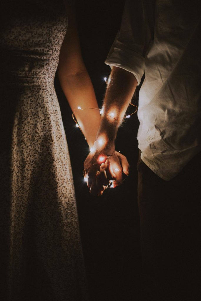 Two people holding hands, with fairy lights loosely wrapped around their hands and forearms.