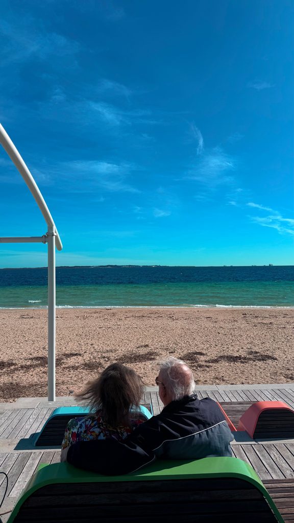 A couple sitting on a bench on a beach, watching the sea